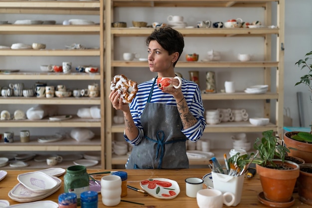 Foto artista di ceramica donna affamata in grembiule che fa colazione mentre è da sola al suo negozio di ceramiche