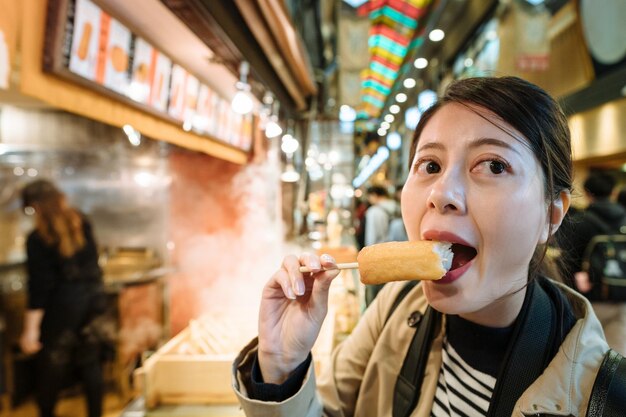 Hungry female tourist opening mouth wide is eating japanese\
traditional food in japan. chinese woman looking aside is having a\
bite on her oden in a local market in kyoto.