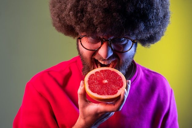 Hungry hipster man with Afro hairstyle biting half of grapefruit, looking at fruit with crazy expression, wearing red sweatshirt. Indoor studio shot isolated on colorful neon light background.