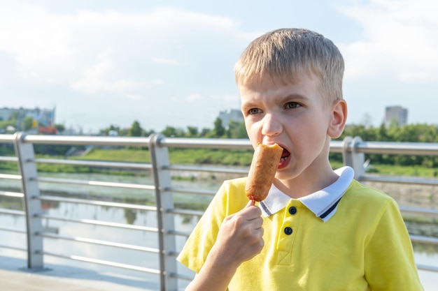 Hungry kid eating street food on the beach in summer A little emotional boy eating fried sausages on a stick