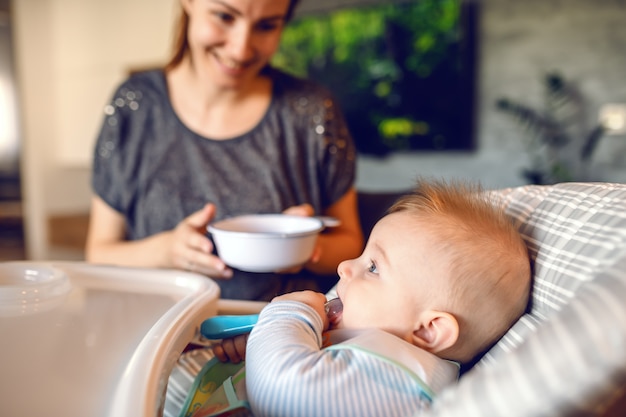 Photo hungry little baby boy with bib sitting in his chair and licking spoon.