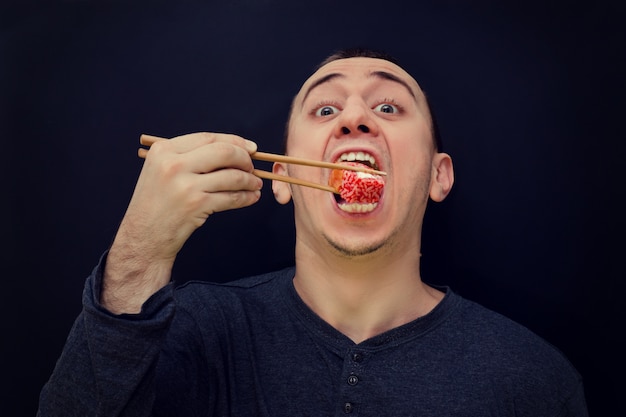 Hungry man eats rolls with chopsticks. Open mouth. Black background