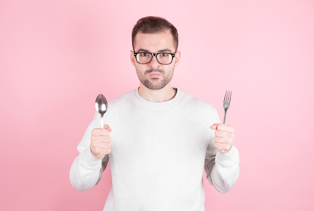 空腹の男は手にフォークとスプーンを持って、ピンクの壁の上のおいしい食べ物について考えます。食事と食事の概念。