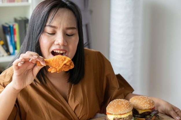 フライドチキン、木の皿にハンバーガー、テーブルにピザを持っている空腹の太りすぎの女性、自宅で仕事中に、体重の問題が発生します。過食症（BED）の概念。