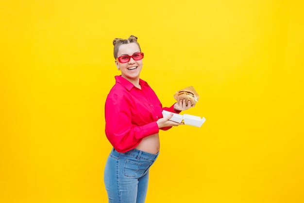 Голодная беременная женщина, держащая гамбургер, ест нездоровую пищу, позирует на желтом фоне в студии Женщина наслаждается большим гамбургером Концепция нездорового питания и переедания во время беременности