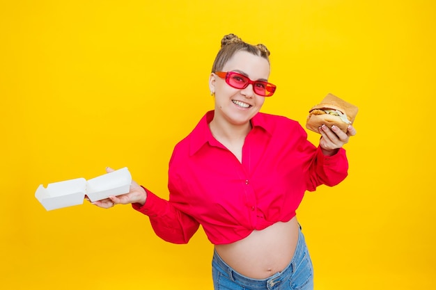 Hungry pregnant woman holding hamburger eating junk food posing on yellow background in studio Woman enjoying a big hamburger The concept of unhealthy eating and overeating during pregnancy