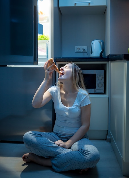 Голодная женщина ест пиццу на полу кухни поздно вечером