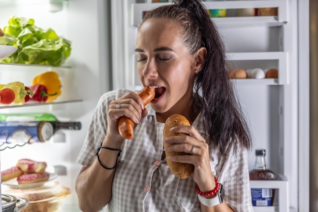Фото Голодная женщина в пижаме ест колбасы с хлебом у холодильника ночью