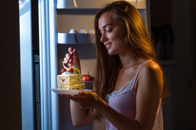 Hungry woman in pajamas eating sweet cake at night near fridge