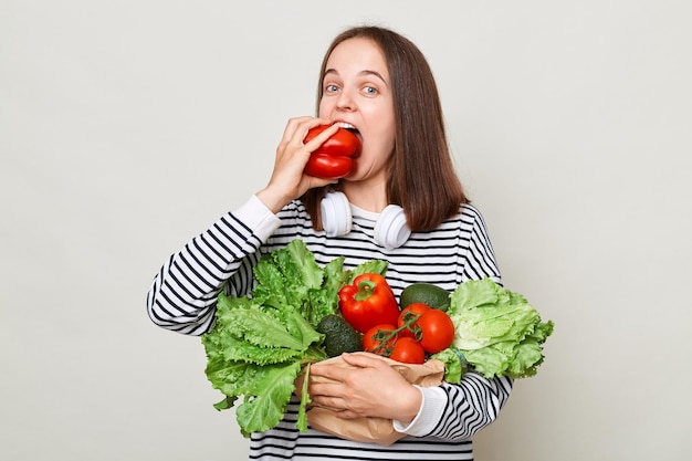 Голодная женщина с каштановыми волосами в полосатой рубашке, стоящая изолированно на сером фоне, кусает сладкий перец, держа свежие сезонные овощи