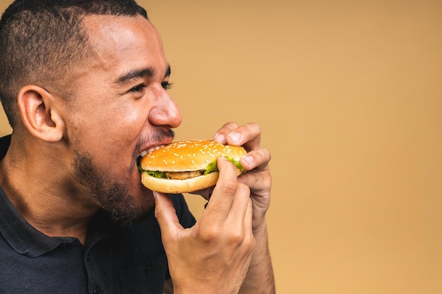 ベージュ色の背景に分離されたハンバーガーを食べる空腹の若いアフリカ系アメリカ人黒人男性ダイエットやファーストフードのコンセプト