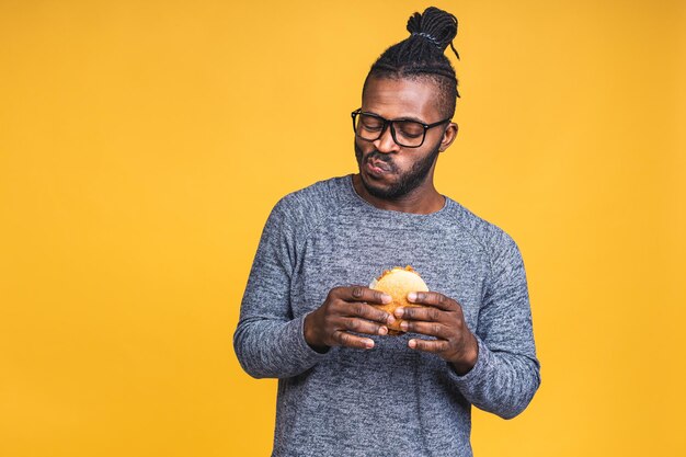 Hungry young african american black man eating hamburger isolated over yellow background. Diet concept.