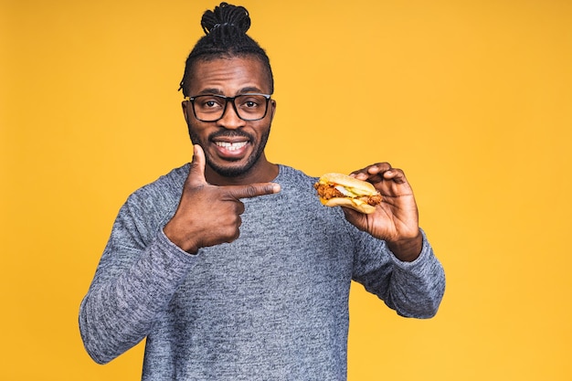 Hungry young african american black man eating hamburger isolated over yellow background. Diet concept.