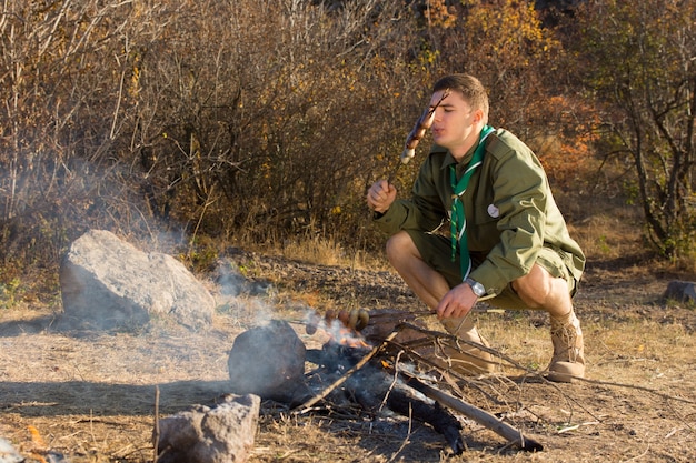 Giovane scout affamato che cucina salsicce su un piccolo fuoco aperto in una radura mentre esplora la natura selvaggia