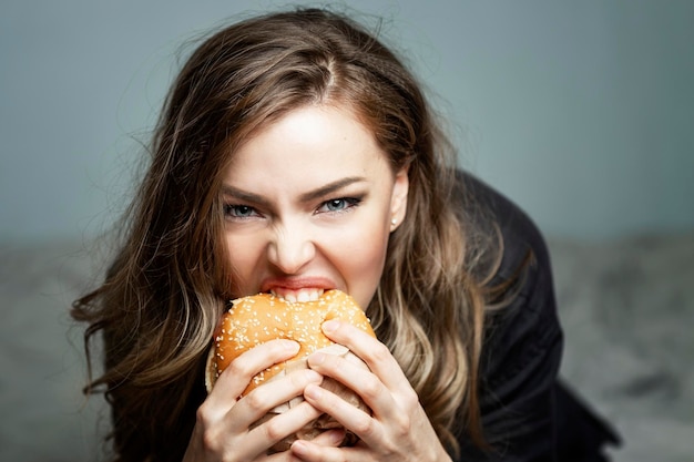 ハンバーガーを食べる空腹の若い女性。美しいブロンド。ジャンクフードと軽食。灰色の背景。閉じる。