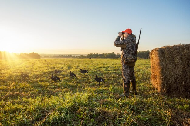 Hunter in camouflage with a gun hunting on black grouse at dawn hunter looking through binoculars at autumn field