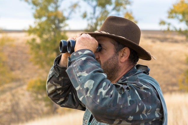 A hunter in a hat with binoculars looks out for prey against the background of the forest