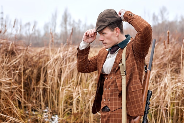 L'uomo del cacciatore regola il cappello in un vestito alla moda il cacciatore con la pistola da caccia in natura guarda il peri di caccia laterale