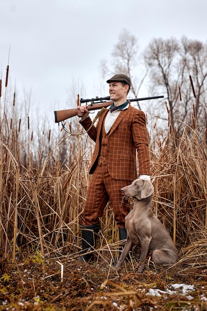 Photo hunter with gray hunting dog in autumn forest. image taken during big game hunting trip. hunting period, autumn season open. handsome caucasian guy in brown suit outdoors in nature