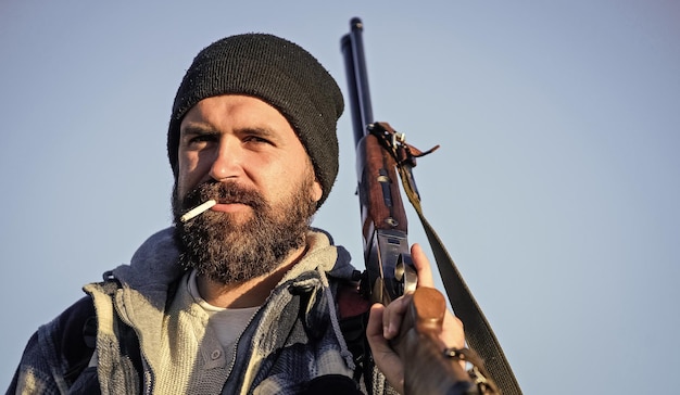 Hunting masculine hobby concept man brutal bearded guy\
gamekeeper blue sky background brutality and masculinity hunter\
with rifle gun close up guy bearded hunter spend leisure hunting\
and smoking