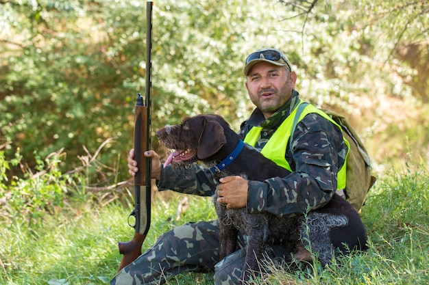 狩猟期間、秋のシーズンが始まります。トロフィーを求めて秋の森で狩猟服を手に銃を持ったハンター。男は武器と狩猟犬と一緒に立ってゲームを追跡します。