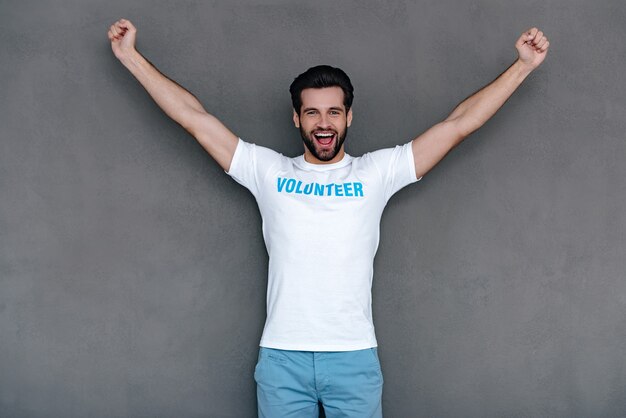 Hurray! Cheerful young man in volunteer t-shirt reaching out his armsand looking at camera with smile while standing against grey background