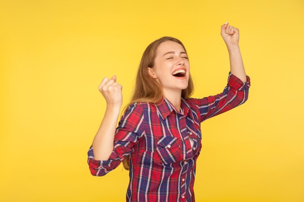 Hurray triumph Portrait of delighted happy girl in checkered shirt rejoicing victory with closed eyes and hands up enjoying success goal achievement studio shot isolated on yellow background