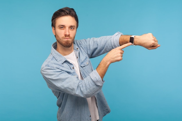 Sbrigati ritratto di uomo arrabbiato impaziente in camicia di jeans da lavoratore che punta all'orologio da polso ed esprime insoddisfazione per la riunione ritardata in ritardo in studio indoor girato isolato su sfondo blu