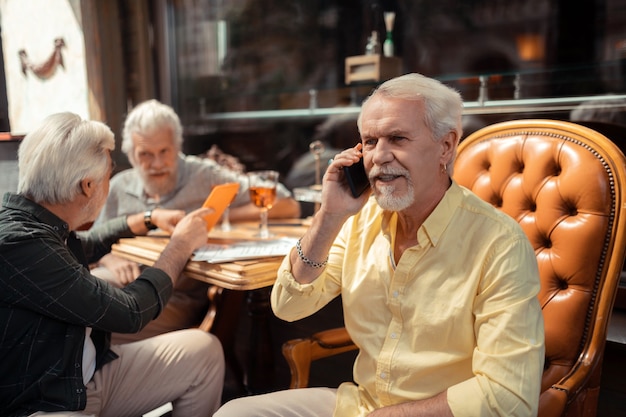 Husband calling. Bearded retired man wearing bracelet calling his wife while spending time with friends