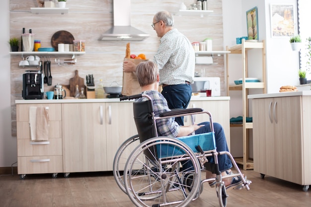 Marito che aiuta la donna anziana disabile in sedia a rotelle con il sacchetto di carta della drogheria. . persone mature con verdure fresche dal mercato. vivere con una persona disabile con disabilità motorie