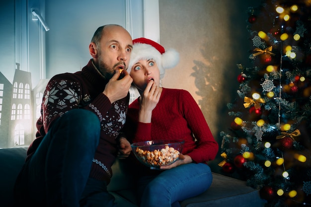 A husband and his wife enjoy popcorn and surprizing while watching TV on Christmas eve