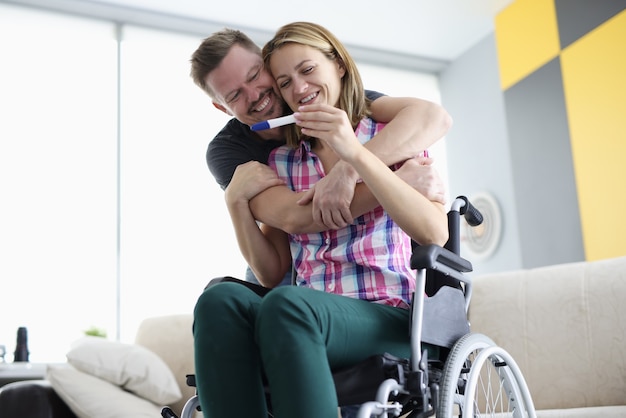 Husband hugs his wife in wheelchair by shoulders with positive pregnancy test