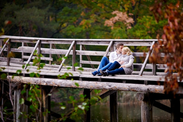 Husband kisses his pregnant wife sitting on a wooden bridge