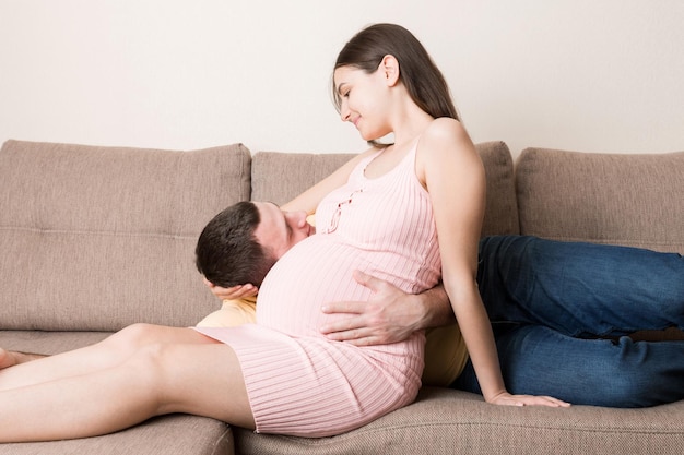 Husband listening to his pregnant wife's belly on sofa at home