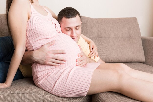 Photo husband listening to his pregnant wifes belly on sofa at home
