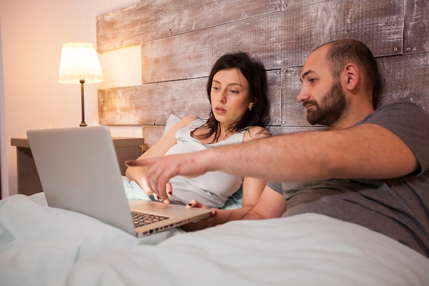 Муж, указывая на ноутбук перед сном. Счастливая супружеская пара в пижаме.