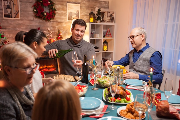 クリスマスのお祝いで義理の父のためにワインを注ぐ夫。幸せな家族