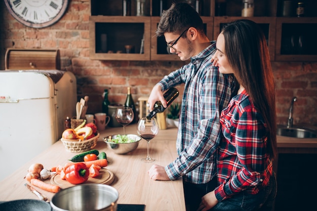 Foto il marito versa il vino in un bicchiere, famiglia che cucina insieme una cena romantica. la coppia prepara insieme la cena romantica. insalata di verdure che cucina sulla cucina. preparazione di cibi dietetici freschi