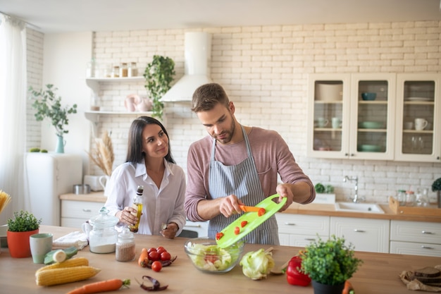 Муж в фартуке добавляет помидоры в салат во время готовки с женой
