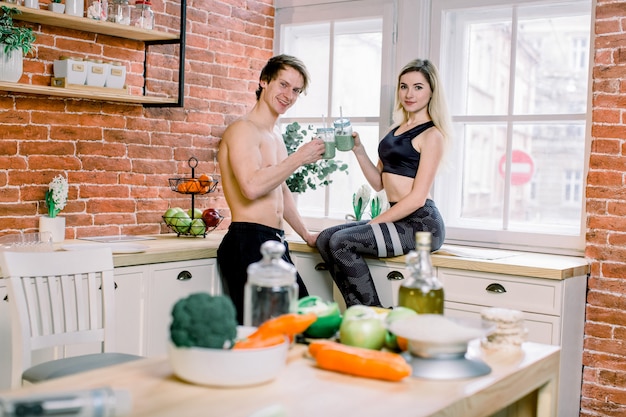 Husband and wife drinkink fresh smoothie in the kitchen and looking at each other. Young sport couple has a breakfast from fresh vegetables and fruits in kitchen at home