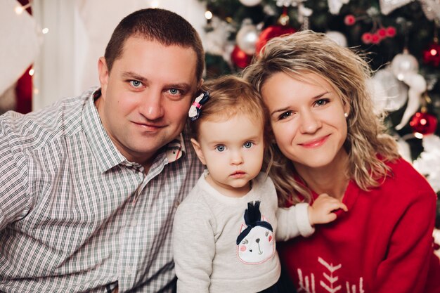 Husband and wife kissing on sofa with little daughter in presents by Christmas tree.