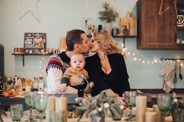 Husband and wife kissing while holding a cute baby and sitting at a festive Christmas dinner table. Holiday concept