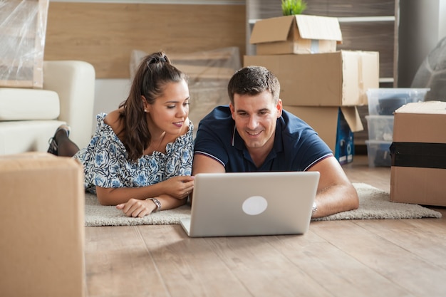 Husband and wife in new appartment lying down on the floor and shopping online on a computer for new furniture