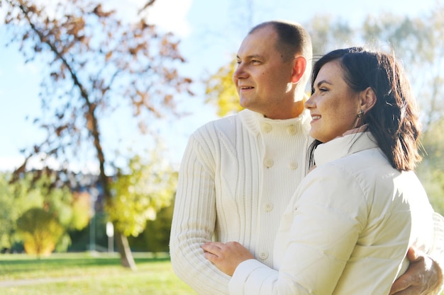 Husband and wife in white warm clothes pose for the camera in an autumn park on a sunny day