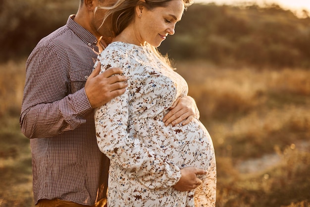 Husband with pregnant wife with big belly tummy is resting on bank of river in summer sunny weather Happy couple in love caucasian man and woman hugging holding hands outdoors