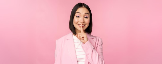 Hush taboe concept portret van aziatische zakenvrouw weergegeven: shush gebaar shhh teken pers vinger aan lippen permanent over roze achtergrond in pak