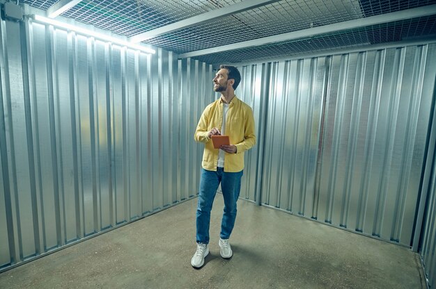 Huur, container. Tevreden man in geel shirt en spijkerbroek met tablet die in een lege ijzeren garage staat en met belangstelling rondkijkt