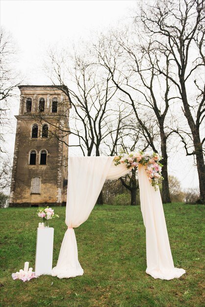 Huwelijksboog versierd met doek en bloemen buitenshuis. Mooie bruiloft opgezet. Huwelijksceremonie op groen gazon in de tuin.