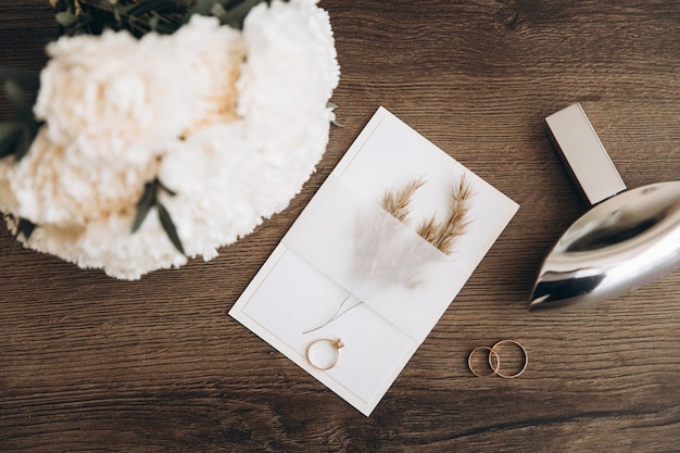 Huwelijksinvitatie of groetekaartje met ambachtelijke enveloppe parfum witte pioenbloemen boeket en trouwringen op houten achtergrond
