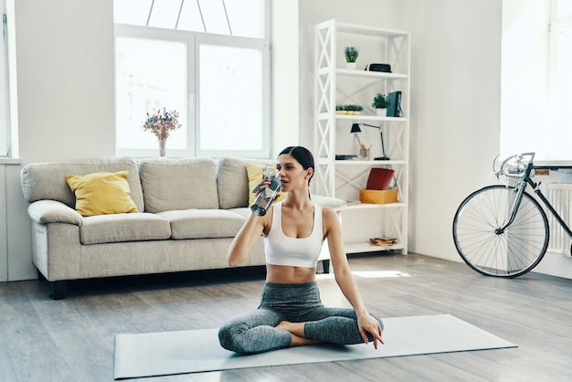 Hydrating. Beautiful young woman in sports clothing practicing yoga and drinking yoga while spending time at home